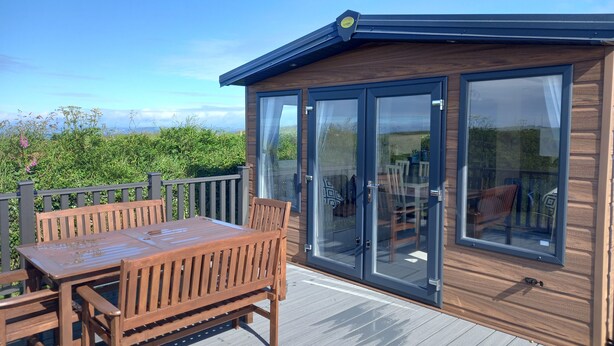 A wooden deck with a table and chairs, a glass door leading to an interior, and a view of greenery in the background. - Valley View -panoramic views of Rame Peninsula (Torpoint)