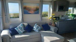A living room with a grey sofa, blue and white patterned cushions, a painting of a beach scene, and a kitchen area with a microwave and a vase of flowers. - Valley View -panoramic views of Rame Peninsula (Torpoint)