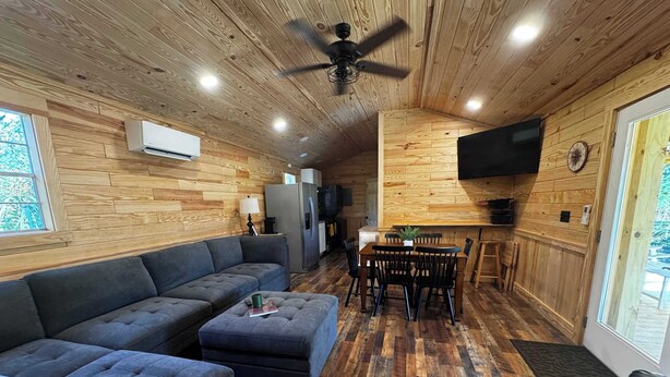 Ein gemütliches Blockhaus-Interieur mit Sofa, Hocker, Esstisch, Stühlen, Deckenventilator und einem an der Wand montierten Fernseher.