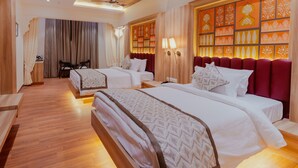 A hotel room with two beds, wooden flooring, a large window with curtains, a seating area with a table, and a decorative wall with patterned panels. - Hotel Ratan Mahal (Datia)