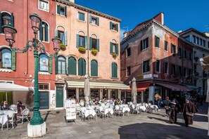 A street with outdoor seating at a café, a green street lamp, and buildings with shutters. - Residenza Santo Stefano (Venice)