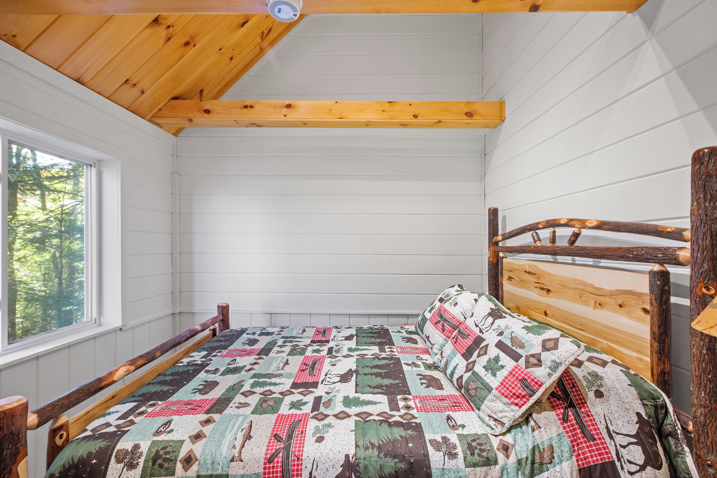 A wooden bed with a quilt in a cabin-style room with a window and white walls.