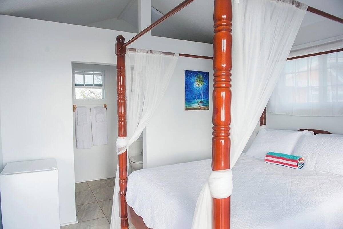 A bedroom with a four-poster bed, a white wall, a painting, and a window with a view.