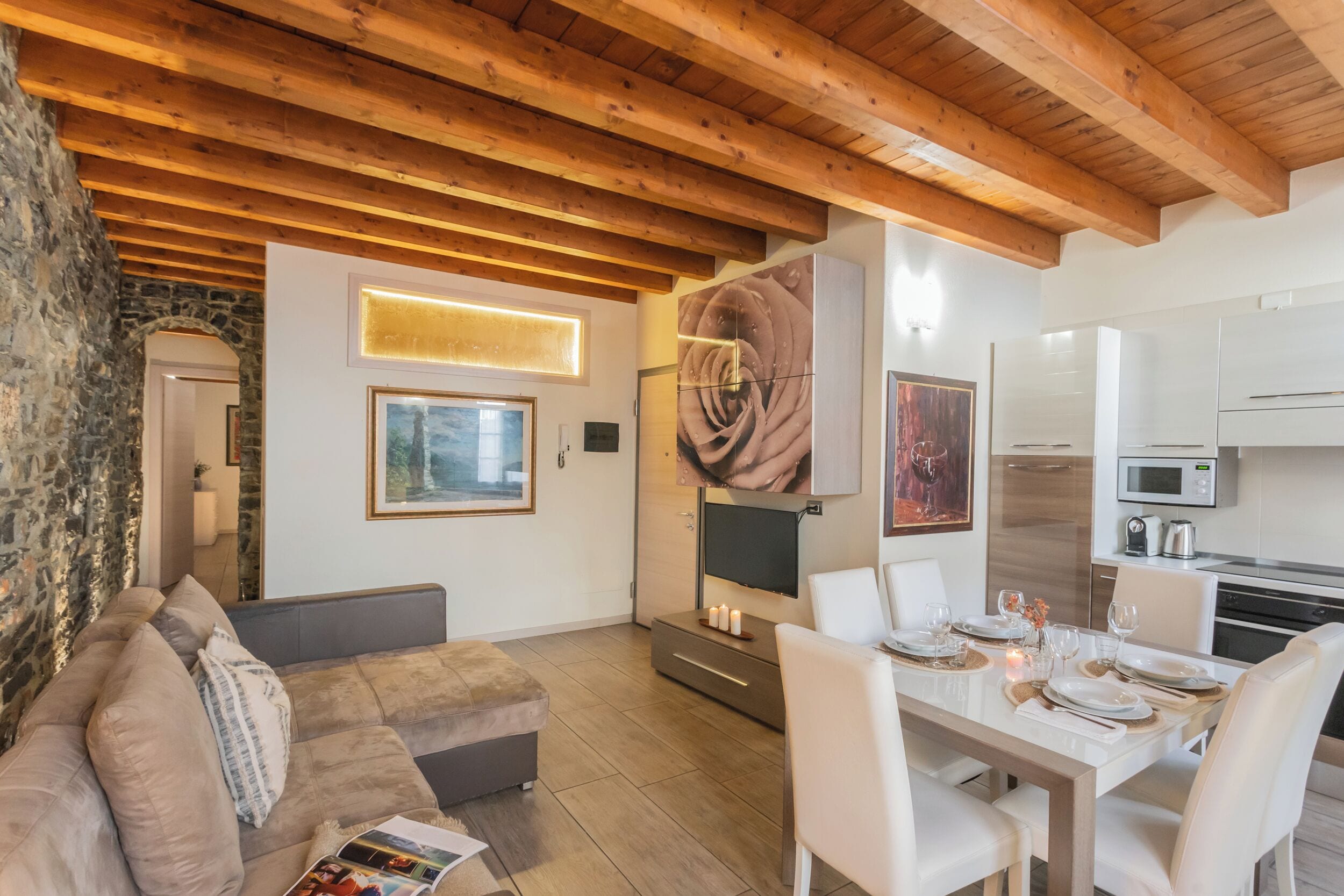 A modern living room with a wooden ceiling, a stone wall, a sofa, a dining table set for four, and a kitchen area with appliances.