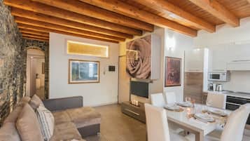 A modern living room with a wooden ceiling, a stone wall, a sofa, a dining table set for four, and a kitchen area with appliances.