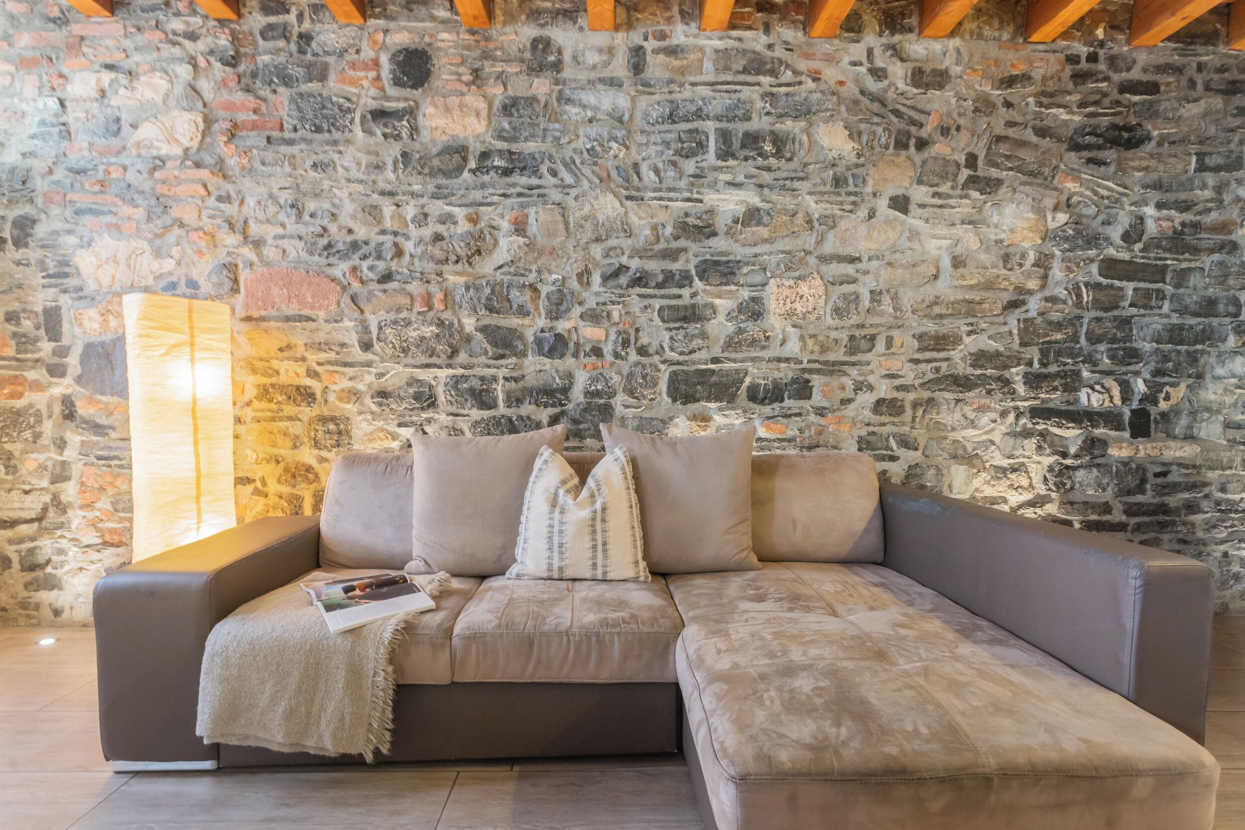 A modern living room with a stone wall backdrop, a grey sectional sofa with a throw blanket, and a magazine on the armrest.