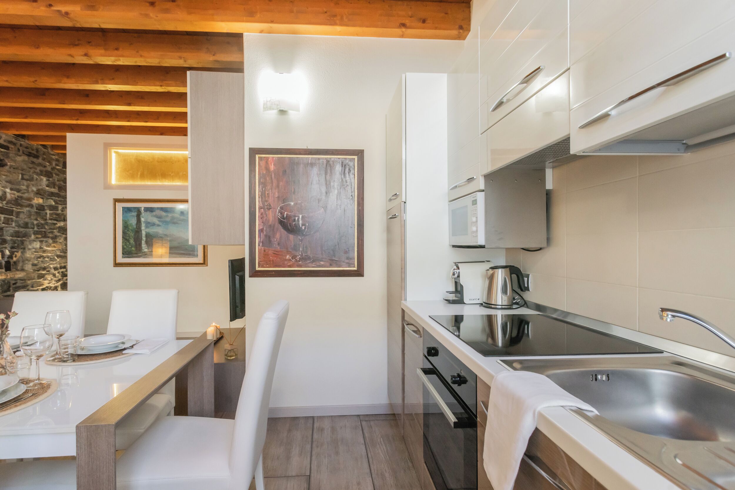 A compact kitchen with white cabinets, a stainless steel sink, and a built-in oven. There is a dining table with chairs and a painting on the wall.