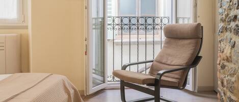 A room with a bed, a chair, a balcony with a metal railing, and a stone wall.