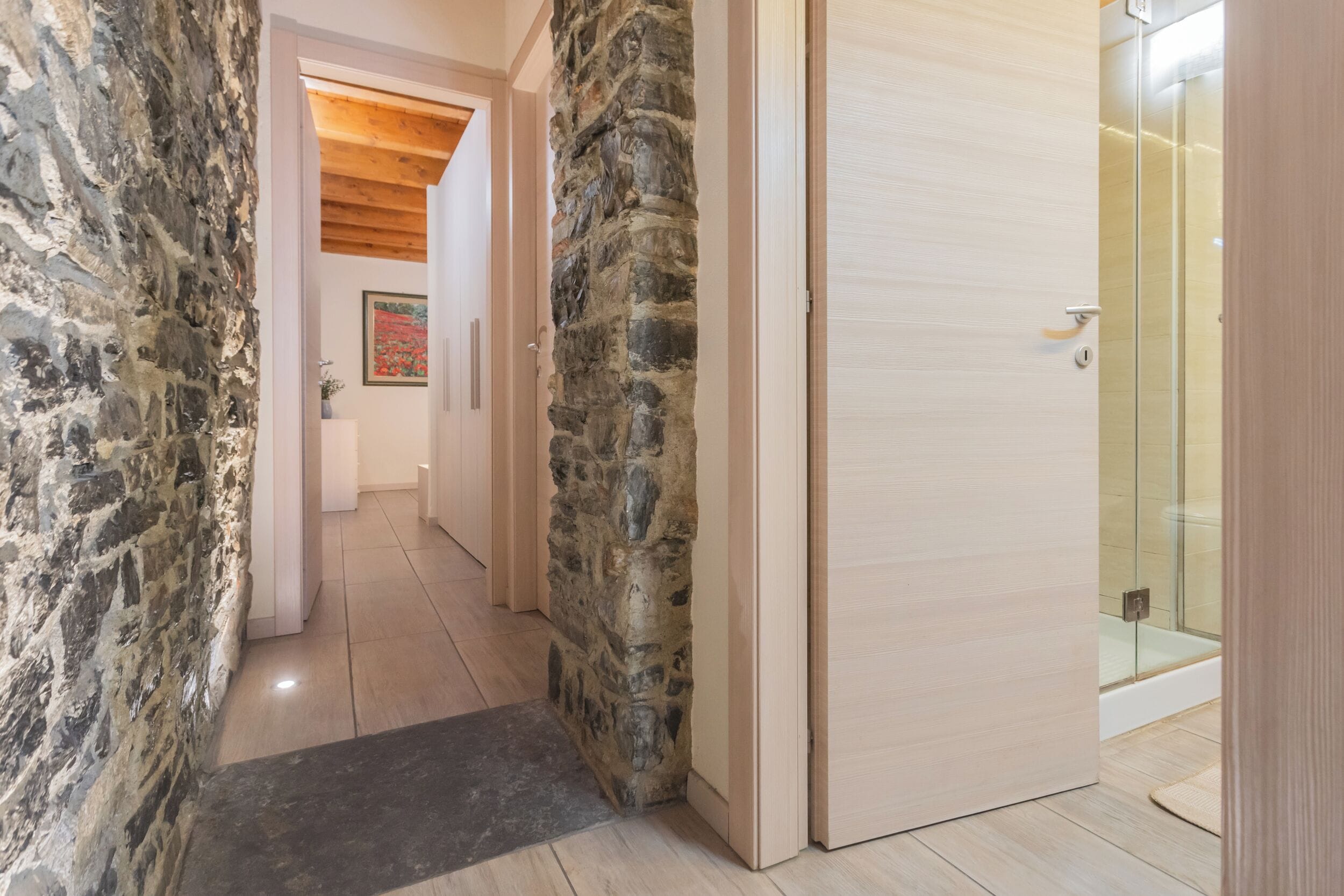 Un pasillo con paredes de piedra, una puerta de madera y una zona de ducha con mampara de vidrio.