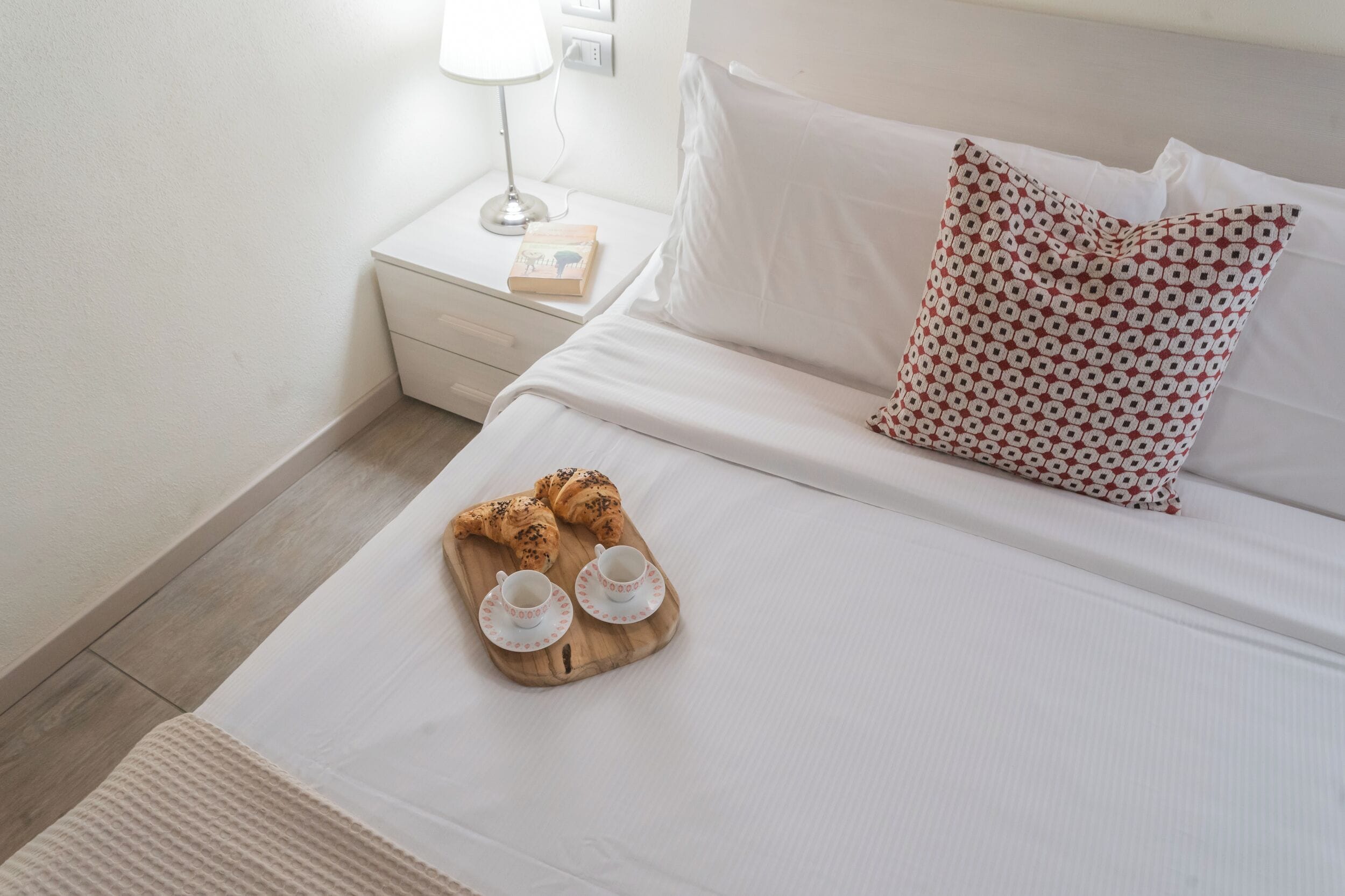 Un lit bien fait, avec des draps blancs, un coussin à motifs et un plateau contenant des croissants et des tasses.
