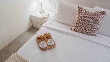 Ein ordentlich bezogenes Bett mit weißen Laken, einem gemusterten Kissen und einem Tablett mit Croissants und Tassen.