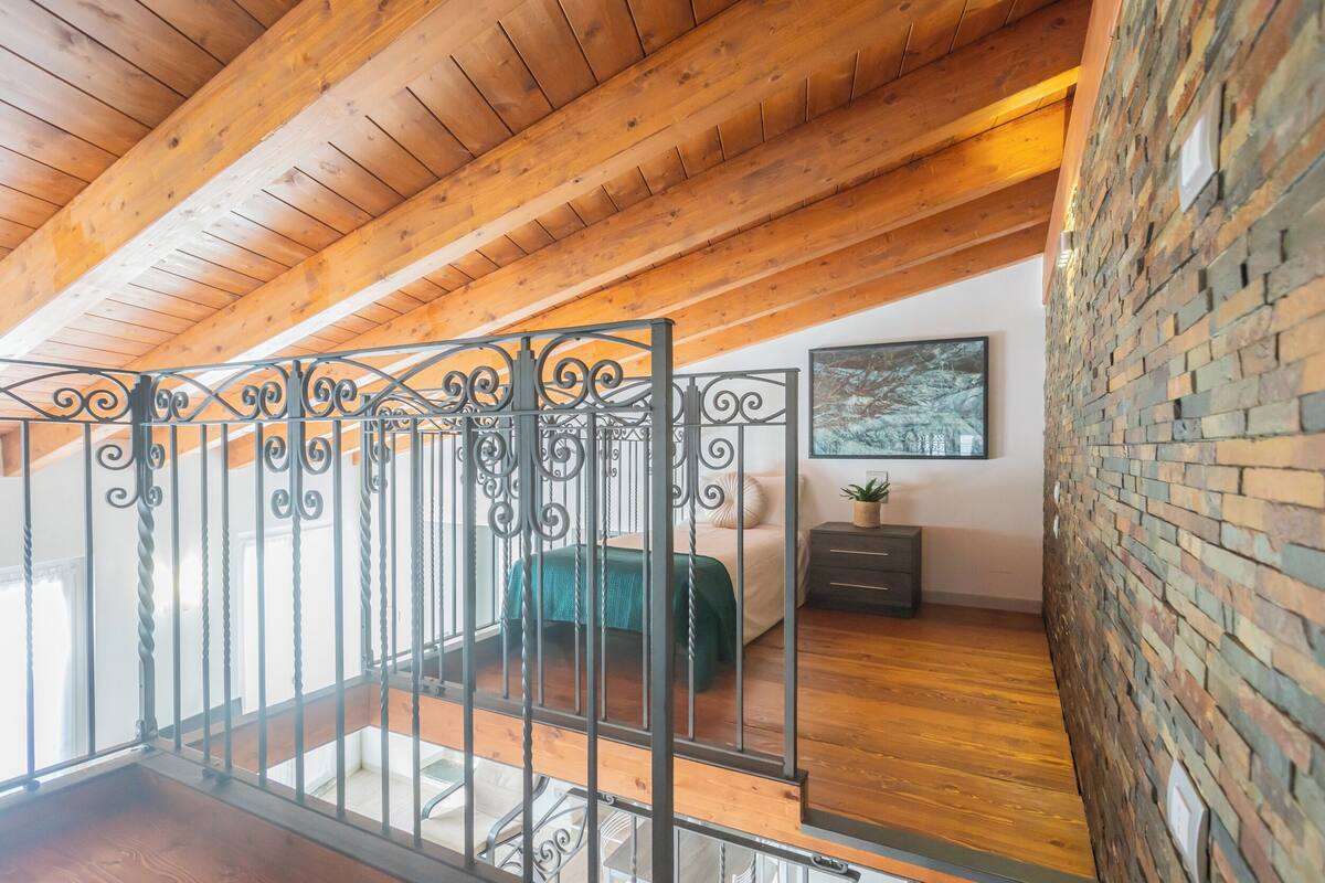 A bedroom with a wooden ceiling, a brick wall, a metal railing, a bed, a nightstand, and a framed picture on the wall.