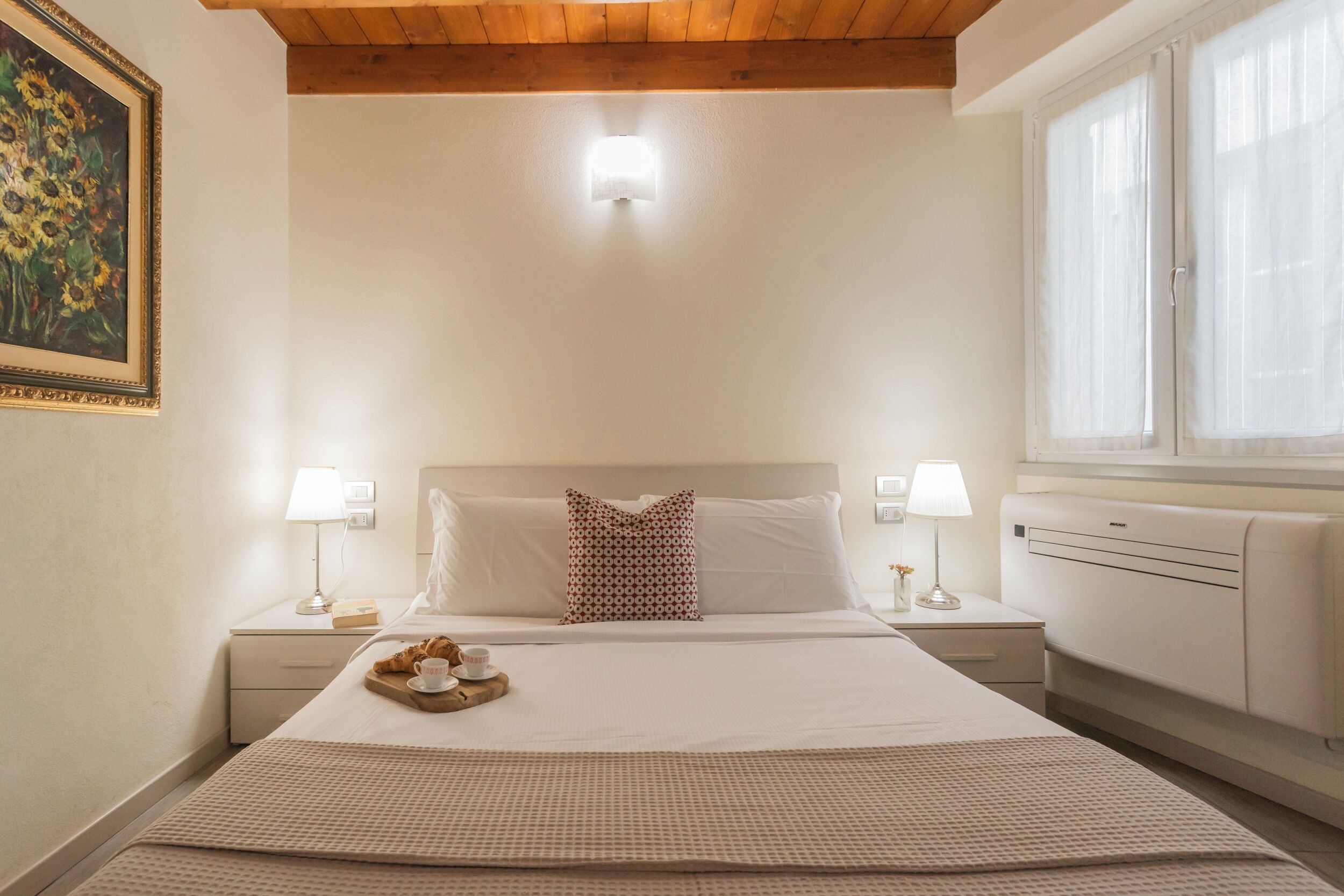 Un dormitorio ordenado con una cama grande, lámparas de noche, una bandeja con croissants y un cuadro en la pared.