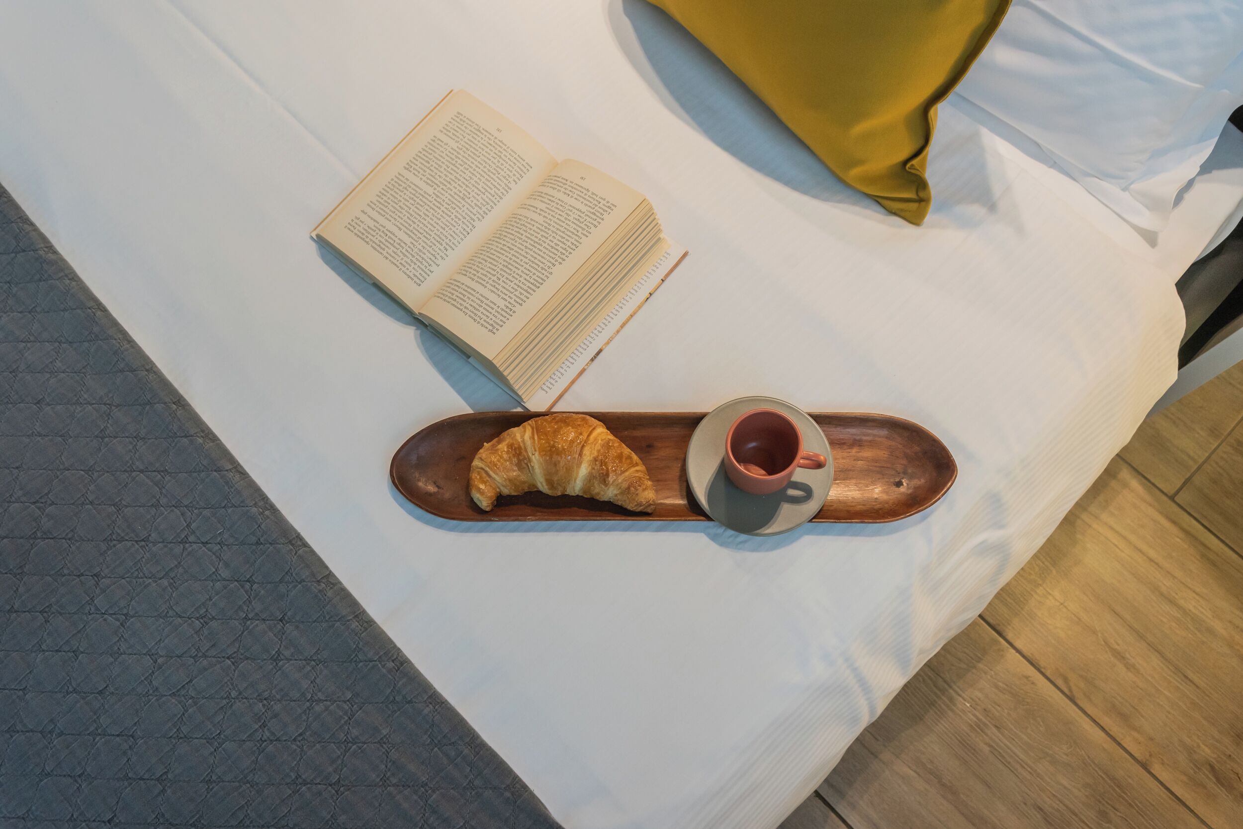 A bed with a book, a croissant, and a cup on a tray.