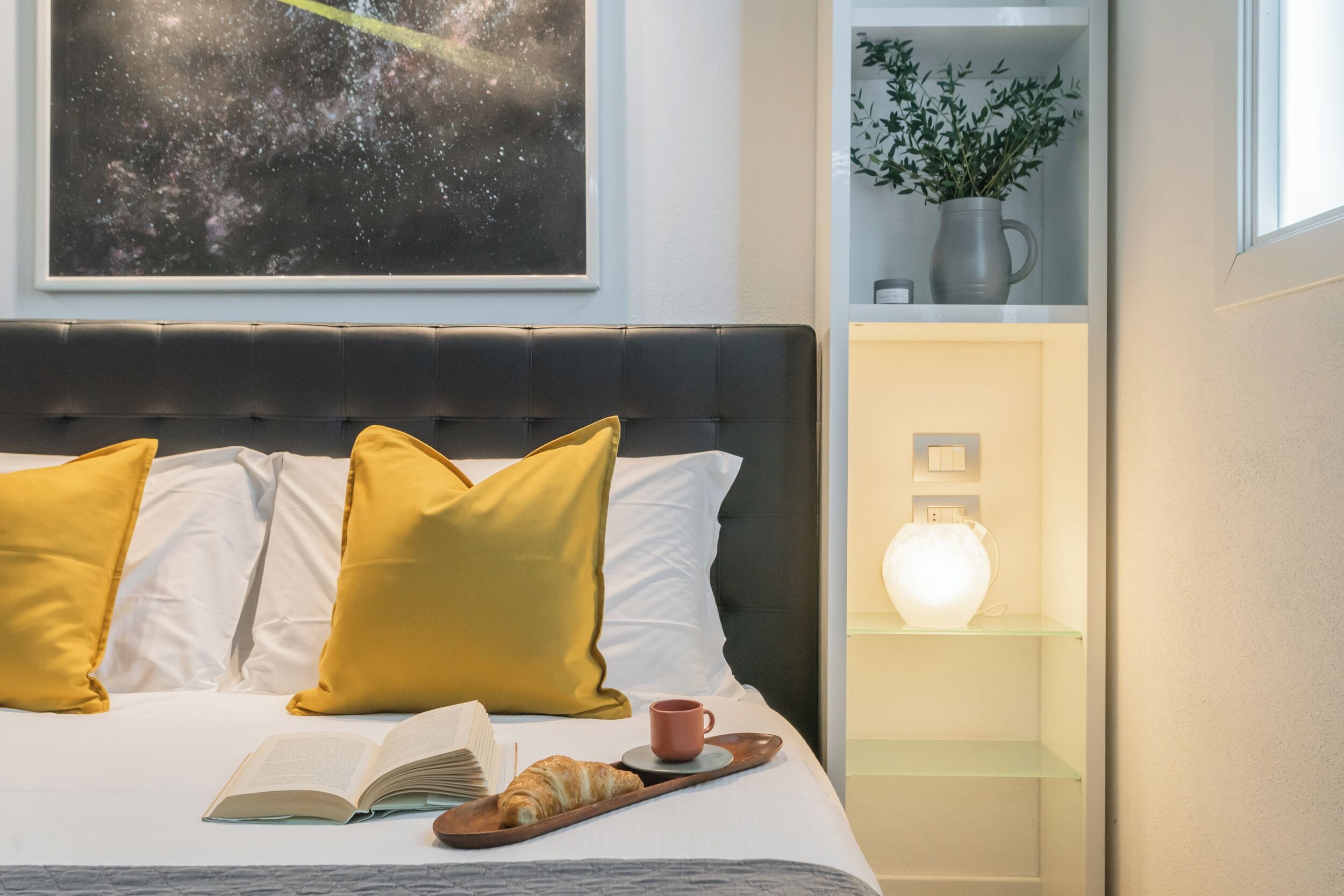 A neatly made bed with yellow pillows, a book, and a croissant on a tray.