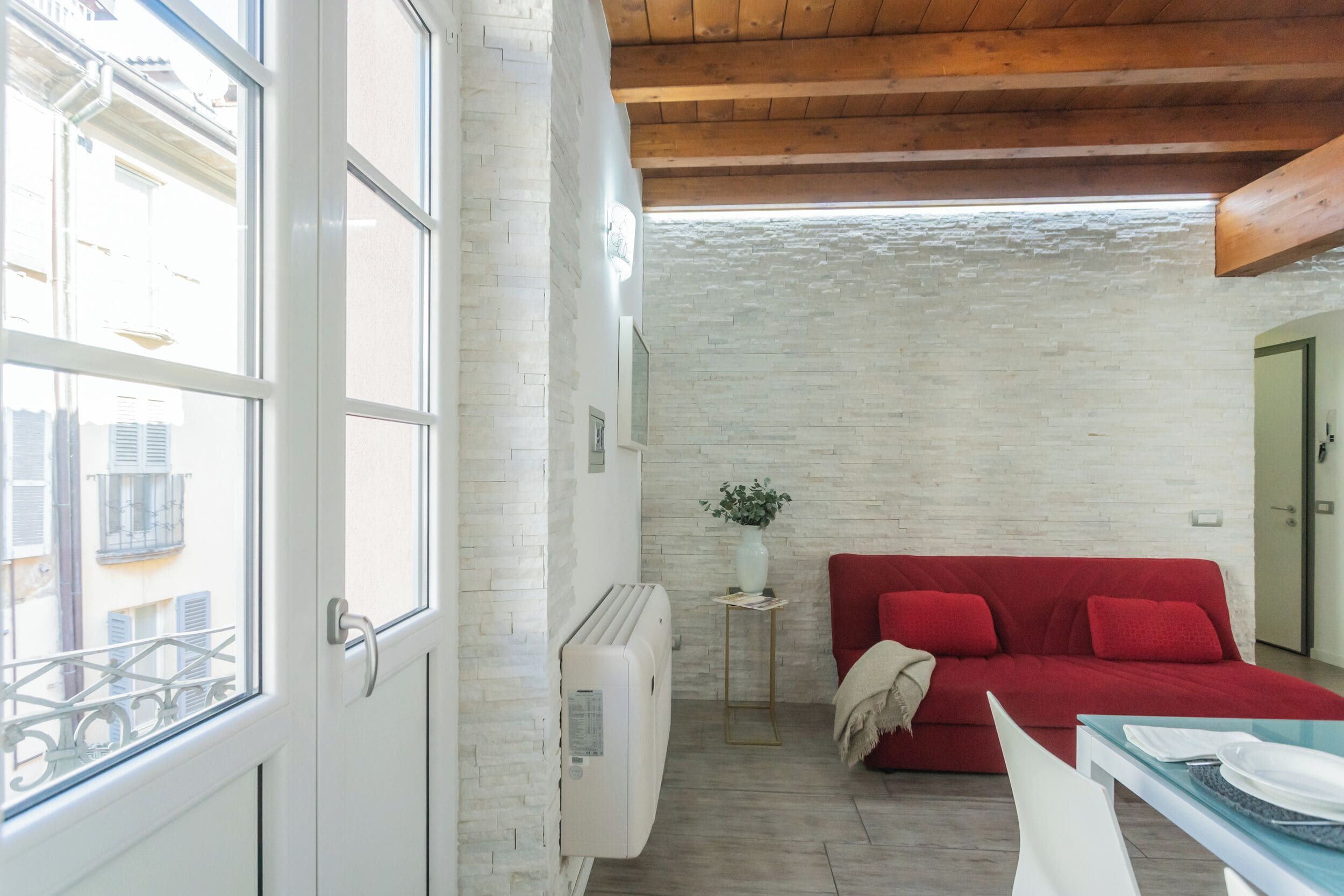 Una habitación con un sofá rojo, una mesita con platos, un radiador blanco y una ventana con vistas a edificios.