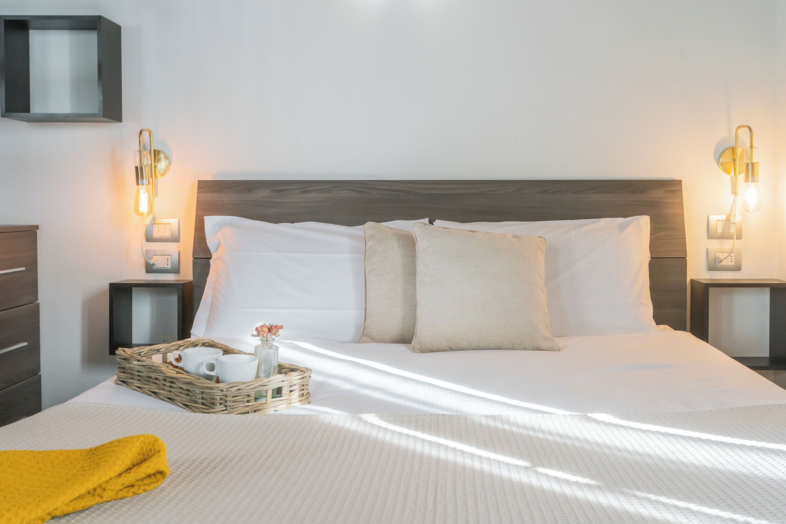 A neatly made bed with white linens, a wooden headboard, and two bedside tables with wall-mounted lamps. A wicker tray with cups and a small vase is on the bed.