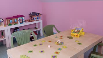 Ein Zimmer mit einem Holztisch, grünen Stühlen und einer rosafarbenen Wand. Auf dem Tisch liegen Spielzeug und eine Spielzeugkiste.