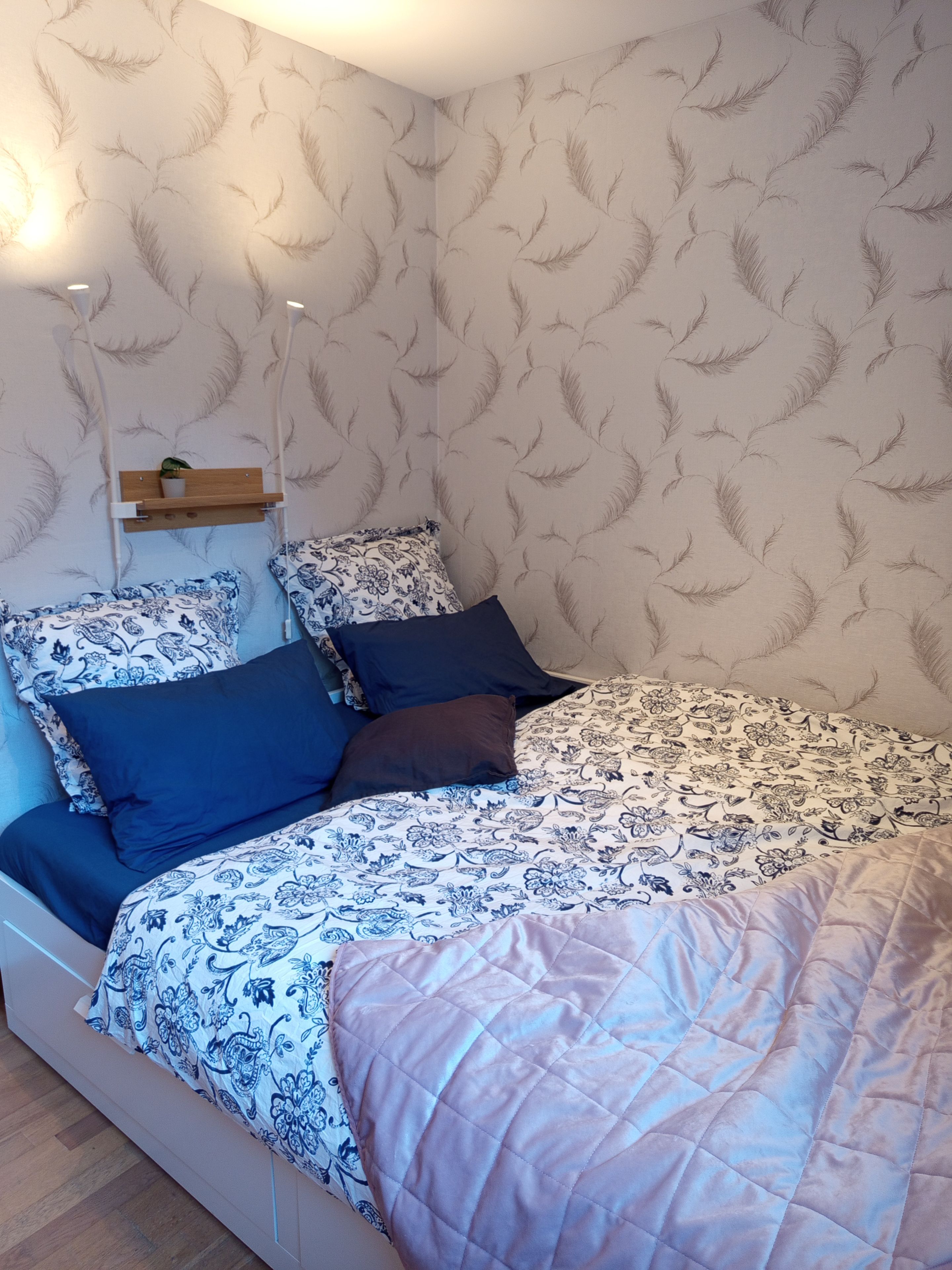 Une chambre à coucher avec un mur à motifs, un lit avec une literie bleue et blanche, et une étagère de chevet ornée d’une plante.