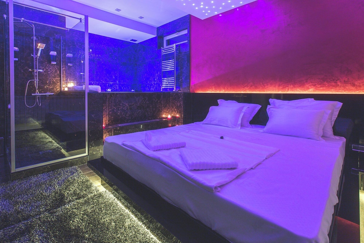 A modern hotel room with a large bed, a glass shower enclosure, and a red accent wall.