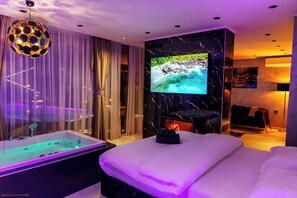 A modern bedroom with a large bed, a bathtub, a TV mounted on the wall, and a fireplace. - Villa Lumiere (Skopje)