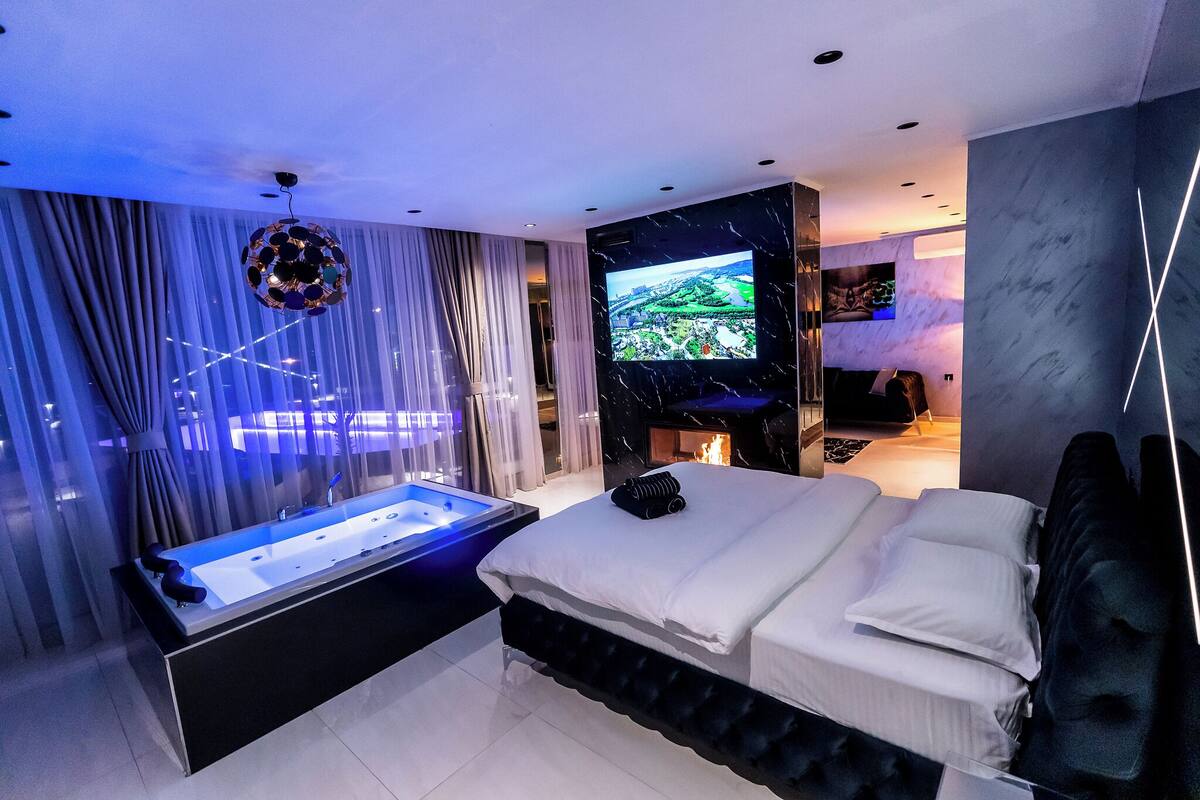 A modern hotel room with a large bed, a bathtub, a flat-screen TV mounted on the wall, and a view of the cityscape through the windows.