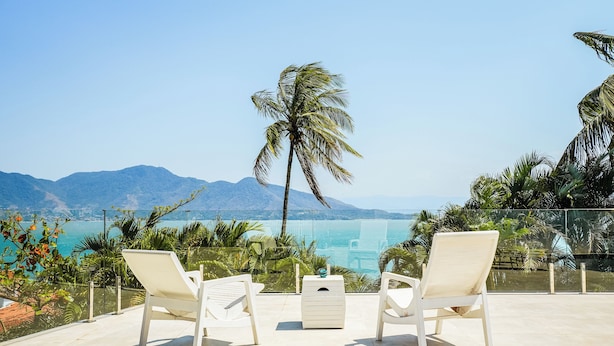 A terrace with two white lounge chairs, a small table, and a view of the sea and mountains. - Casa da Imperatriz (Ilhabela)