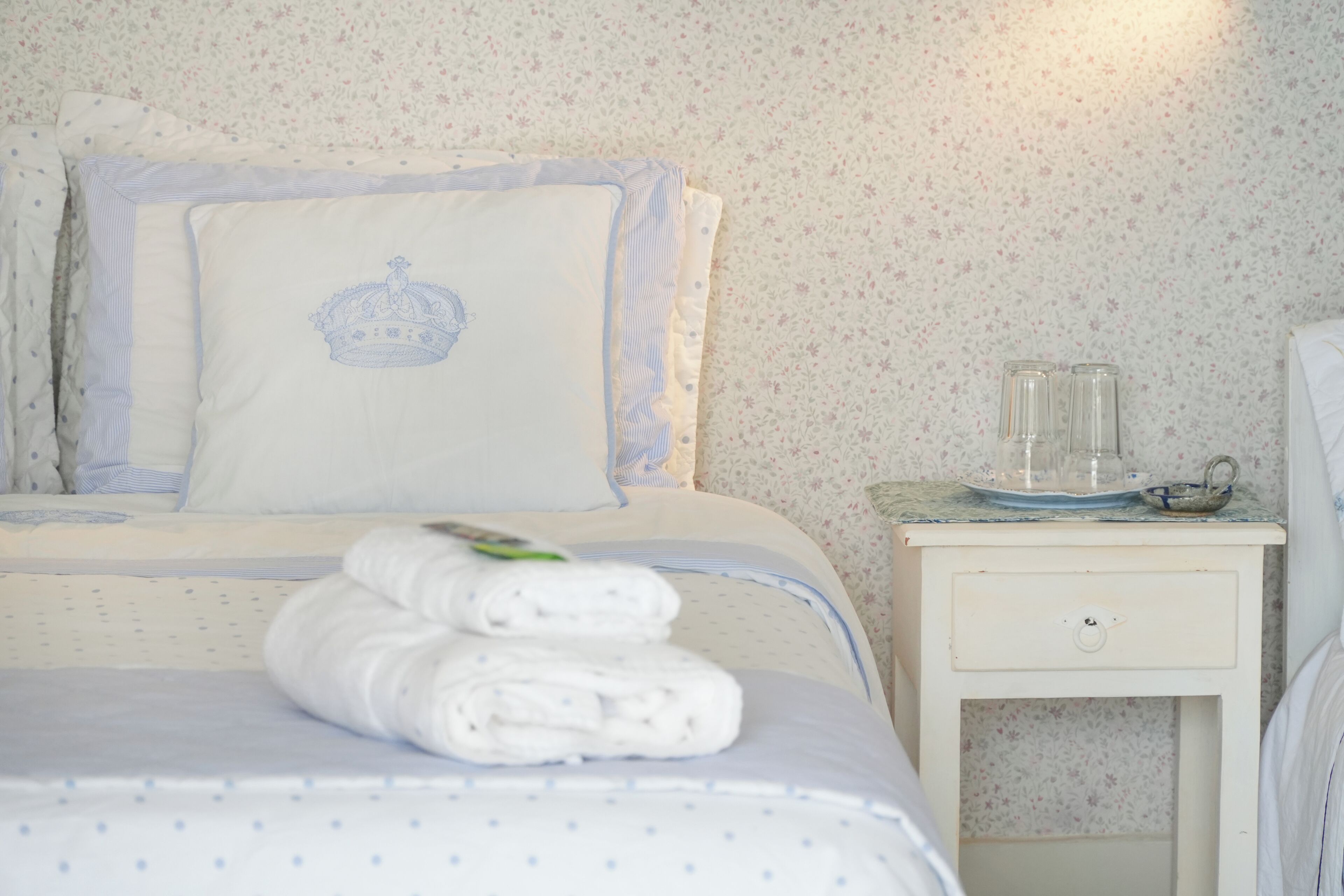 A neatly made bed with a pillow featuring a crown design, a set of folded towels, and a bedside table with a glass container and a bowl.