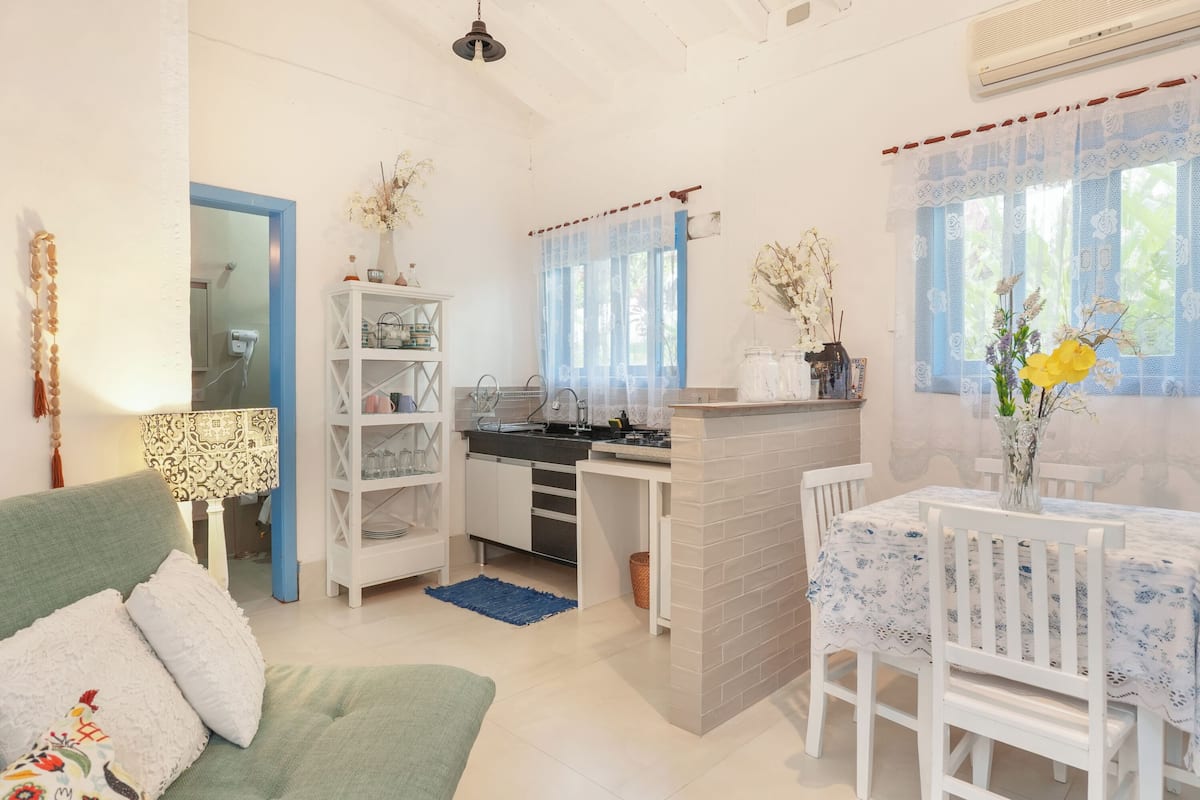 Un salon chaleureux avec un canapé vert, une table à manger avec des chaises blanches et un coin cuisine avec une fenêtre bleue.