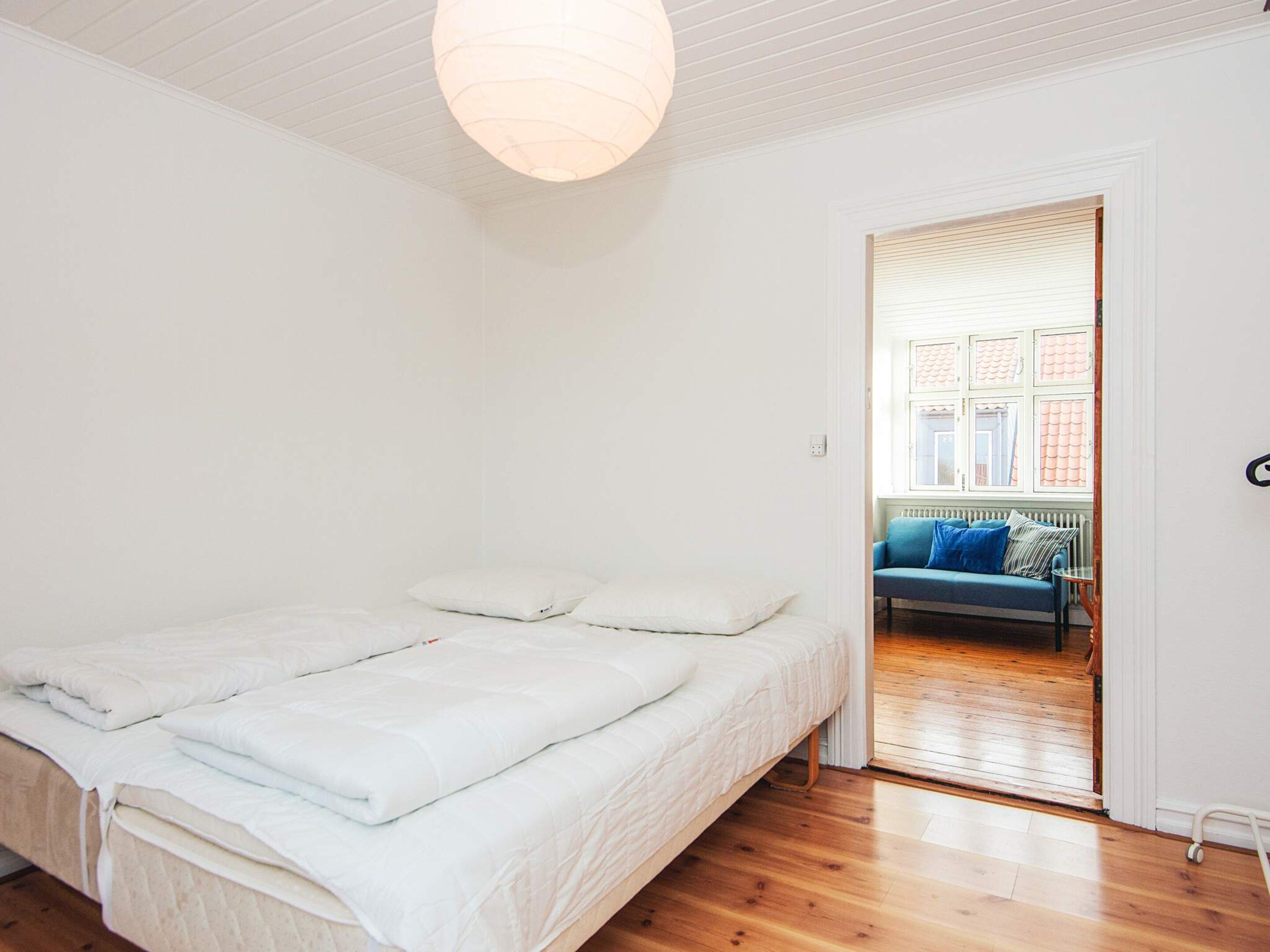 A bedroom with a bed, white walls, a wooden floor, and a view of a blue sofa in the adjacent room.