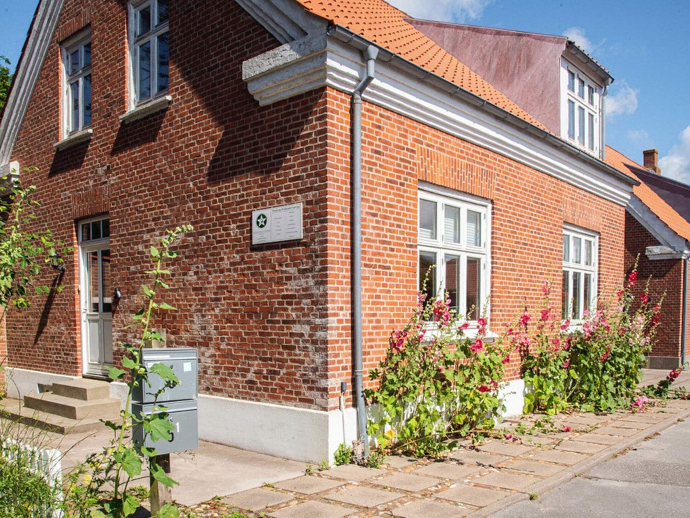 A red brick building with a white window frame, a small garden with pink flowers, and a metal utility box.