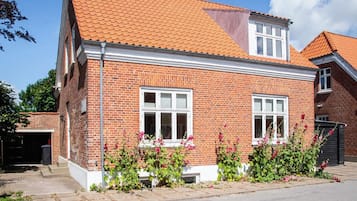 A two-story brick house with a red-tiled roof, white-framed windows, and a small porch area with flowering plants.