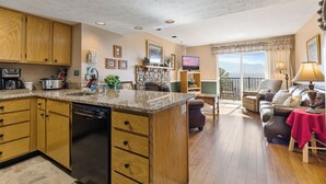Cocina amplia con armarios de madera, lavavajillas y encimera de granito. Cuenta con un comedor con mesa y sillas, y una sala de estar con sofá y televisor. La habitación tiene un ventanal con vistas a la montaña.