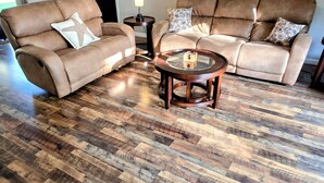 A living room with a brown leather sectional sofa, a glass coffee table, and a herringbone hardwood floor.