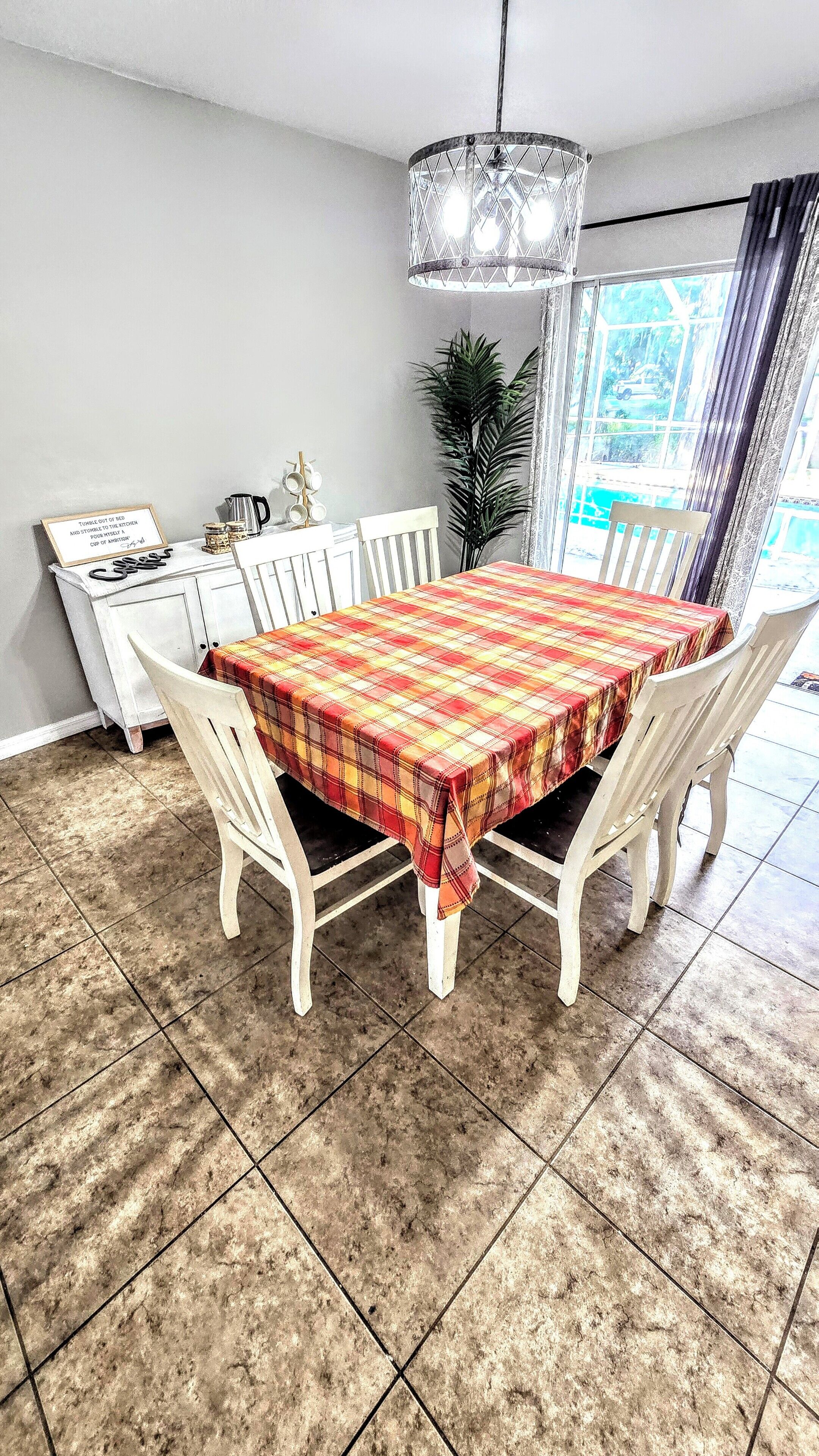 A dining area with a table covered by a plaid tablecloth, surrounded by white chairs, a chandelier, and a plant.