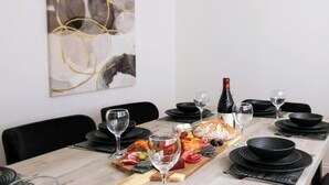 Una mesa de comedor dispuesta con platos, vasos, una botella de vino y diversas opciones de comida, incluyendo pan, queso y embutidos.