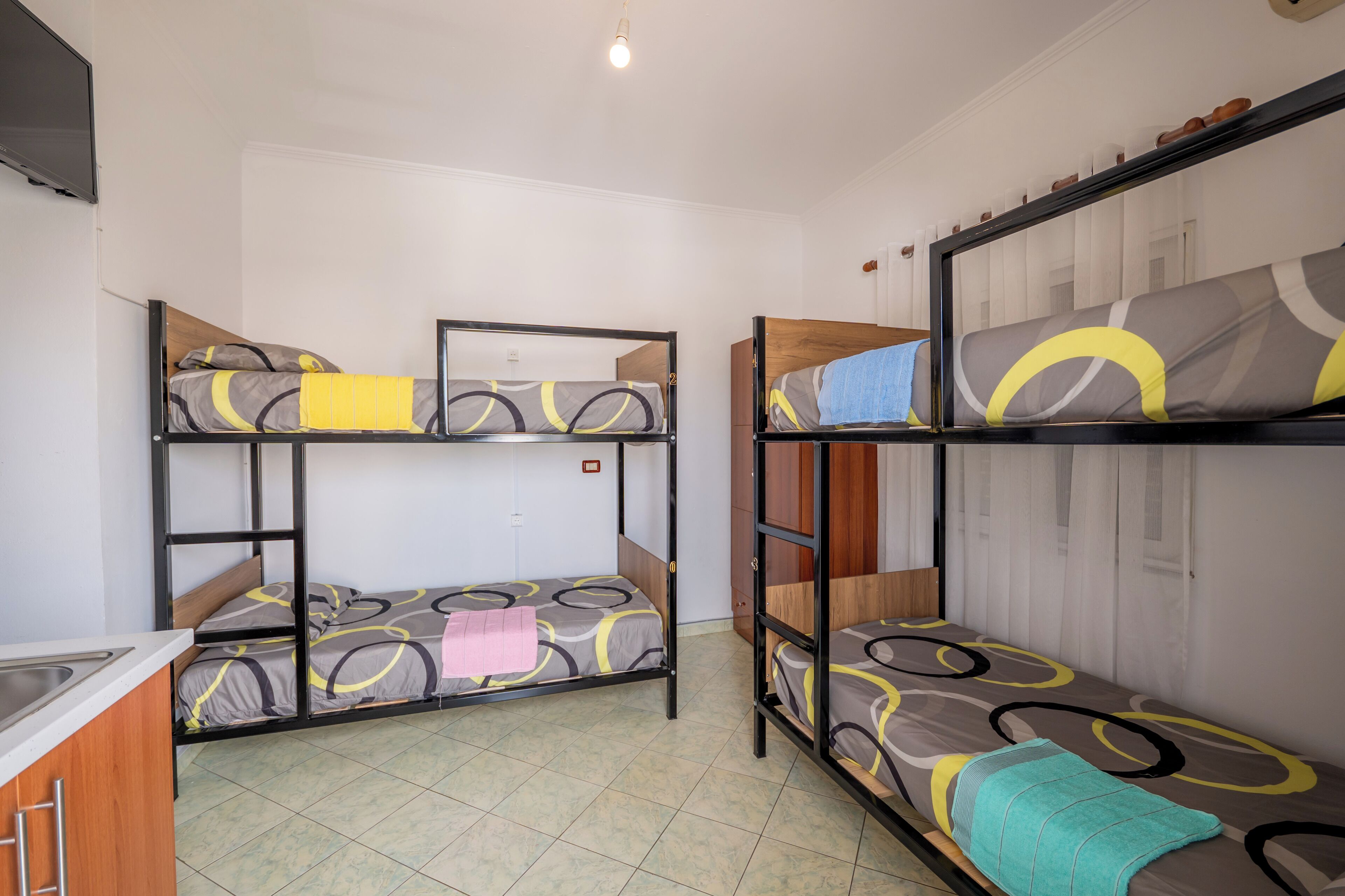 A dormitory room with bunk beds, a kitchen area, and a window with curtains.