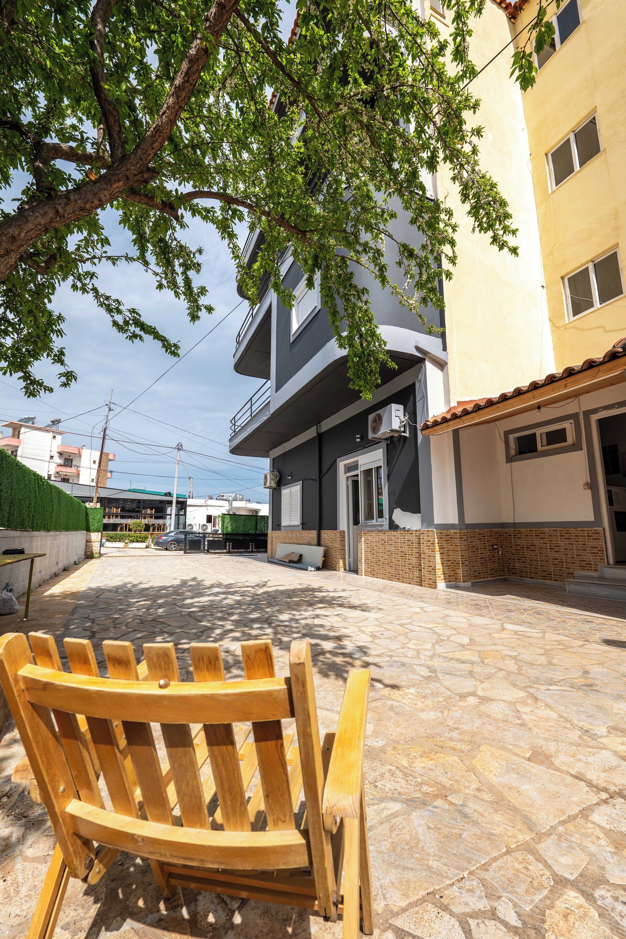 Un patio con un banco de madera, un edificio con balcón y un árbol que da sombra.