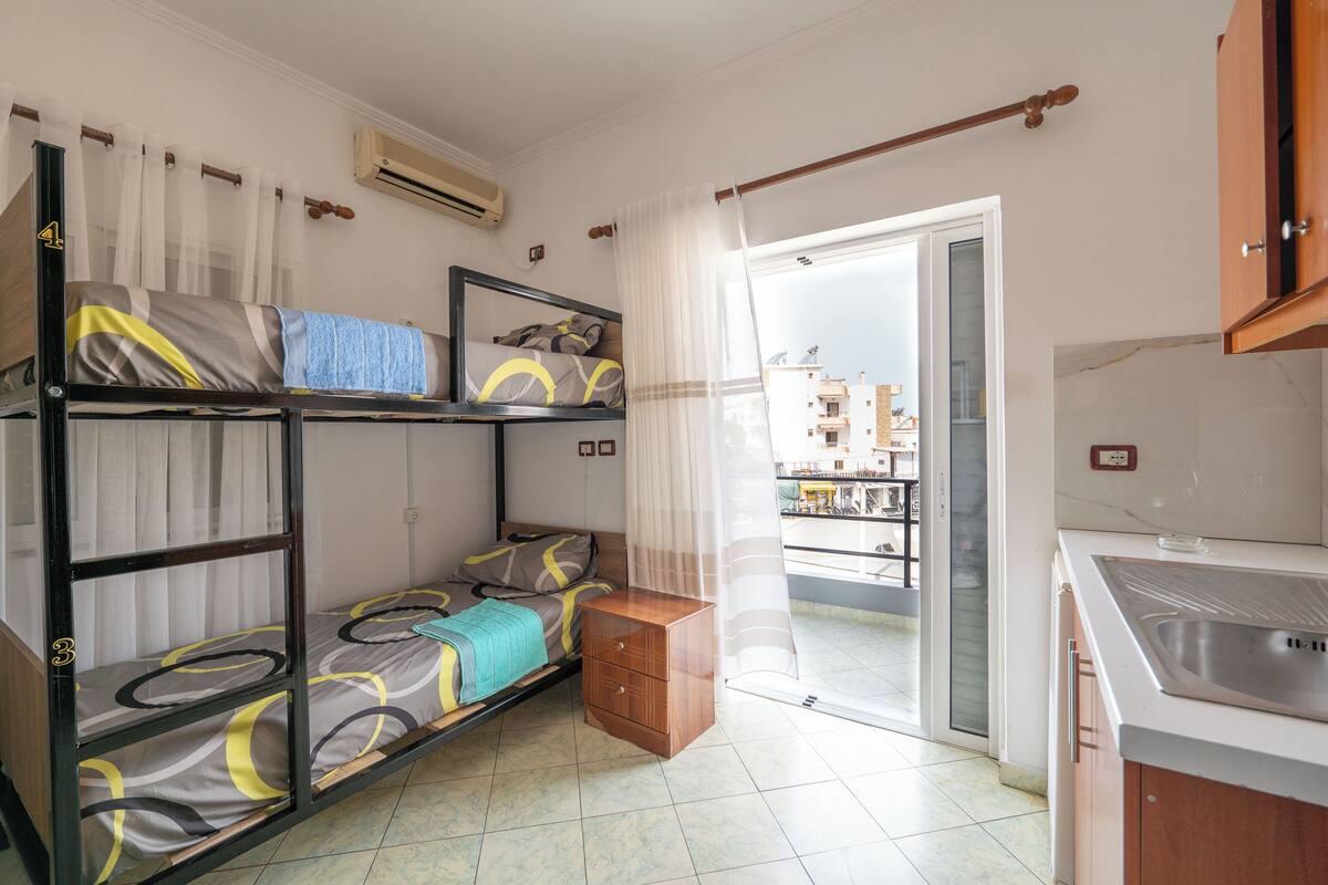 A dorm-style room with bunk beds, a small wooden nightstand, a kitchen area with a sink, and a sliding glass door leading to a balcony.