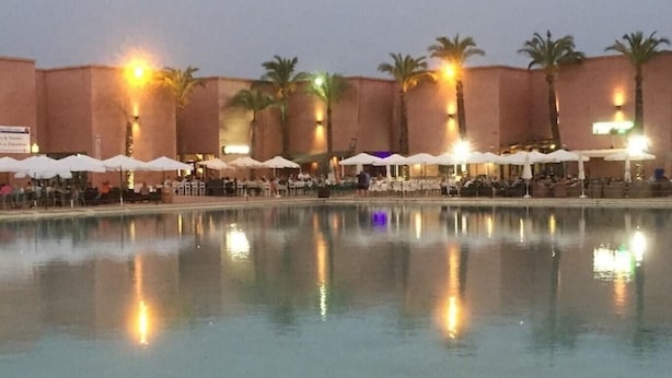 A hotel pool area with multiple umbrellas and palm trees, surrounded by buildings with warm lighting. - Sunny Oasis Retreat in Condado del Alhama (Alhama de Murcia)