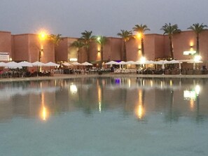 A hotel pool area with multiple umbrellas and palm trees, surrounded by buildings with warm lighting.