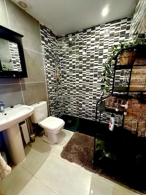 A bathroom with a white sink, toilet, and shower. The walls are tiled with a black and white checkered pattern. There is a mirror above the sink and a shelf with plants and toiletries. - Sunny Oasis Retreat in Condado del Alhama (Alhama de Murcia)