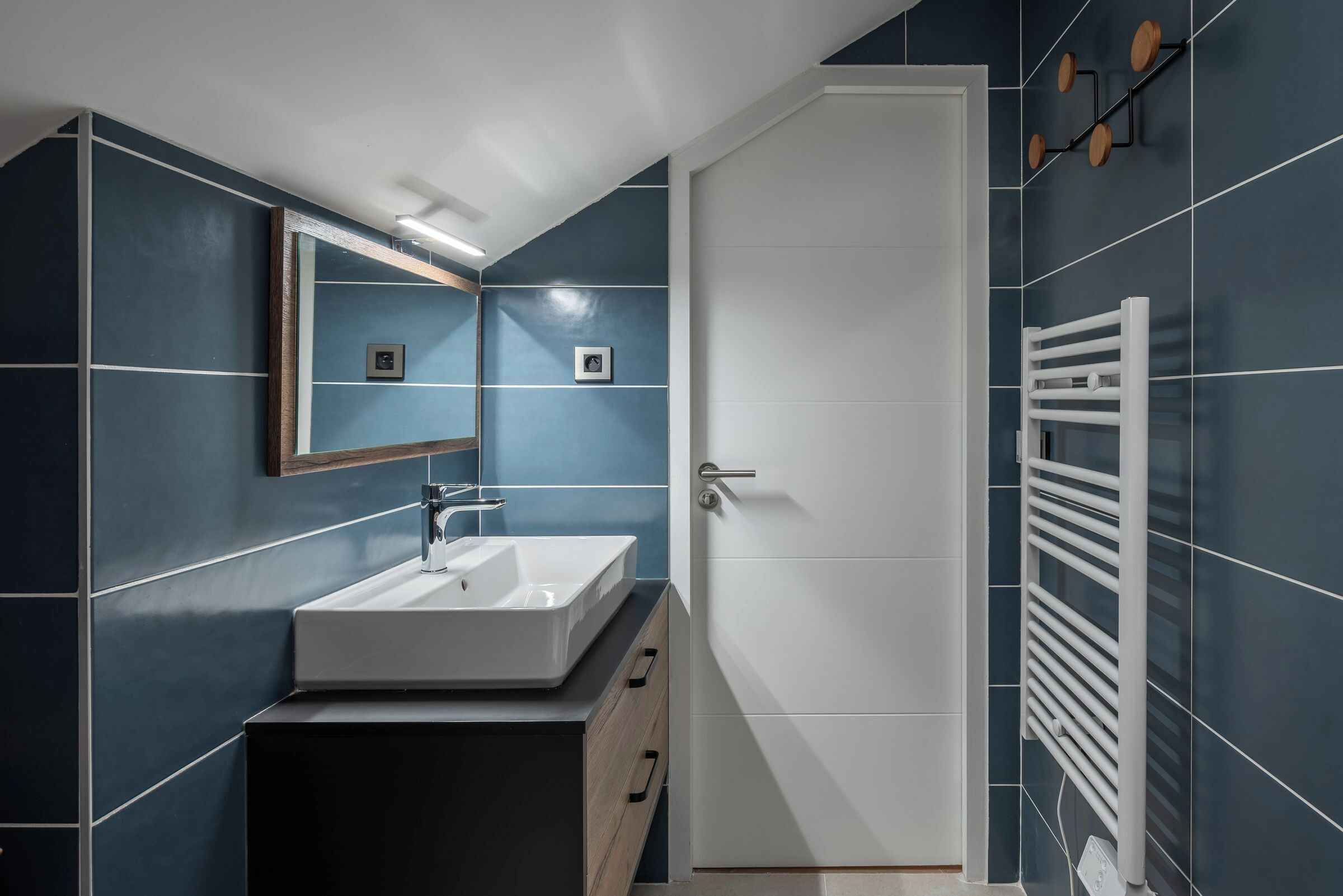 A modern bathroom with a white sink, dark blue tiles, a white door, and a towel rack.