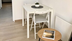 A compact living space with a dining table, a small round table with a magazine, a framed picture on the wall, and a view into another room with a bed. - La Vie Douce - Between Ocean & Marshes (Saint-Michel-en-l'Herm)