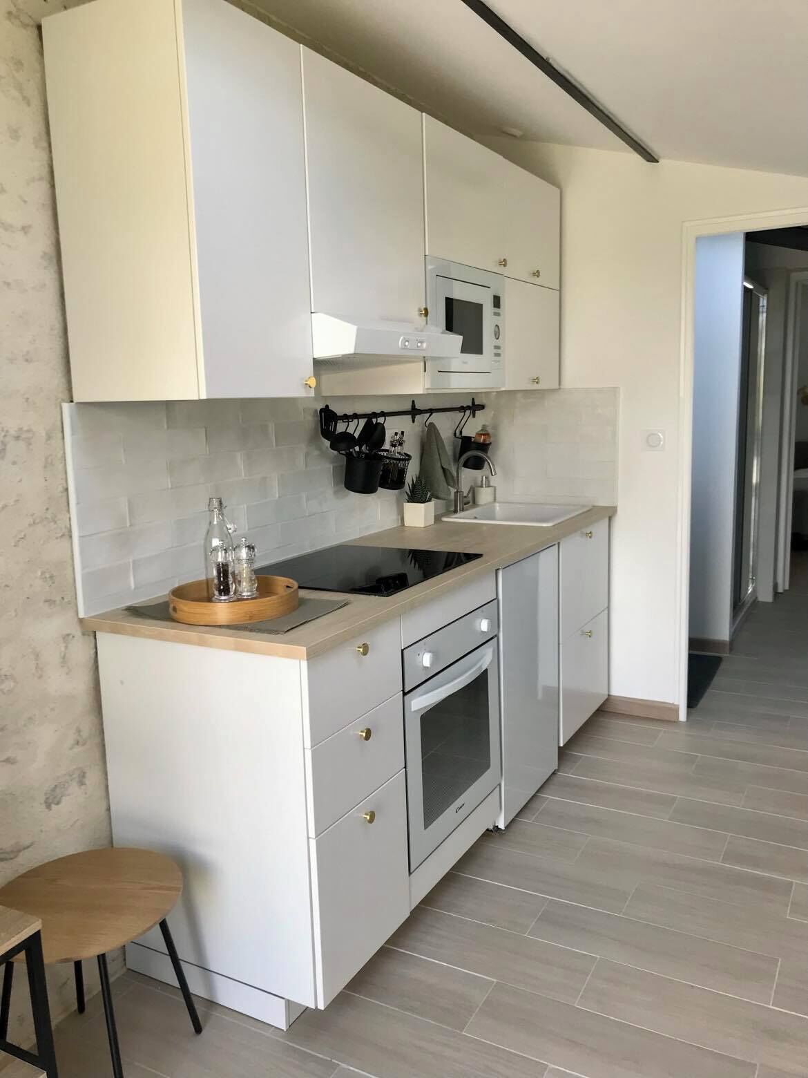 Una cocina moderna con armarios blancos, un microondas, una estufa y una encimera de madera. Hay un taburete pequeño y un colgador para ollas.