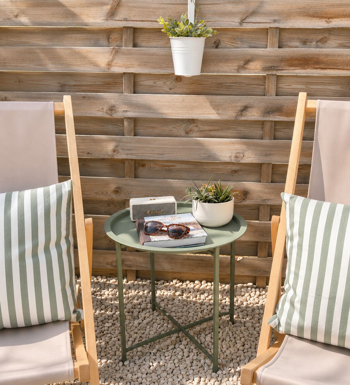 A wooden fence, two striped chairs, a small table with a plant, a book, and sunglasses.