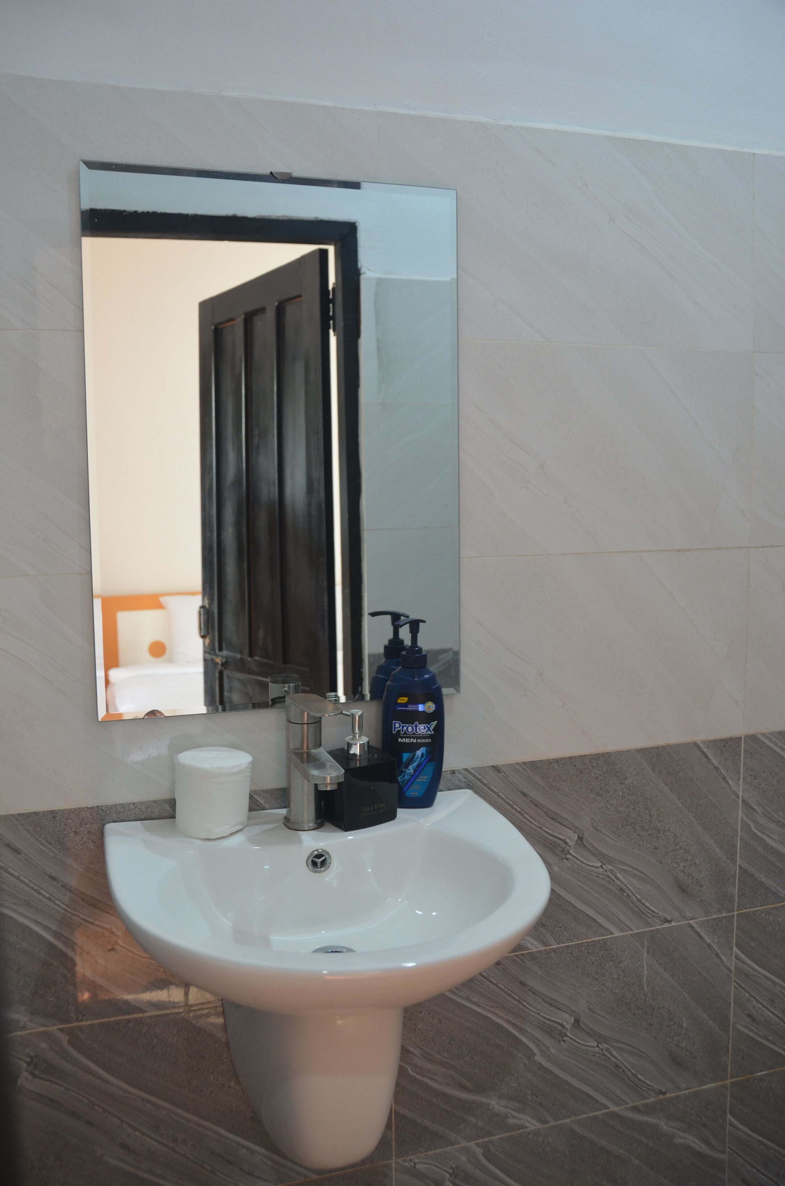 A bathroom with a white sink, a mirror, a bottle of hand soap, and a roll of toilet paper.