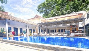 A swimming pool with lounge chairs, a covered area, and a building with a tiled roof.