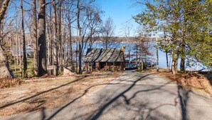 A cabin with a wooden exterior, a dirt path leading to it, and a lake in the background. - New to Lake Norman! Cozy Cabin with Big Water Views! (Sherrills Ford)