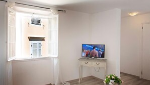 Ein Zimmer mit Fernseher auf einem weißen Konsolentisch, einem Fenster mit weißen Vorhängen, einer weißen Tür und einer Zimmerpflanze auf dem Boden.
